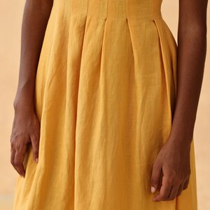 Close-up detail shot focusing on the waistline of a mustard yellow linen dress, showcasing structured box pleats and a side pocket.