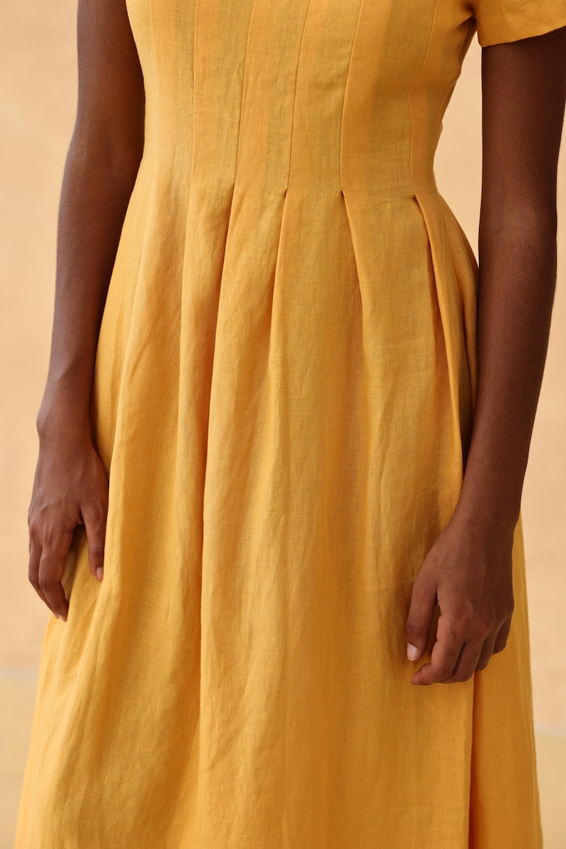 Close-up detail shot focusing on the waistline of a mustard yellow linen dress, showcasing structured box pleats and a side pocket.