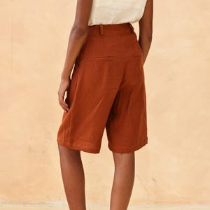 Close-up rear view of the model's lower torso, highlighting the clean tailoring, pocket layout, and structure of the terracotta brown linen shorts or trousers, overlaid with interface editing icons.