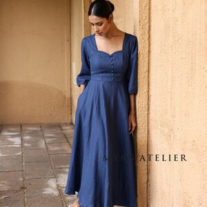 Full-length front shot of a woman with a poised stance walking through a stone arched hallway, showcasing the timeless feminine tailoring of a slate blue linen button-front dress.