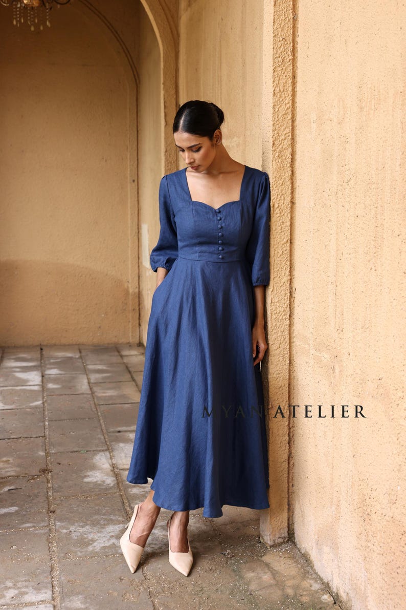 Full-length front shot of a woman with a poised stance walking through a stone arched hallway, showcasing the timeless feminine tailoring of a slate blue linen button-front dress.