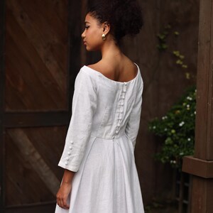 A close-up rear view of the model focusing on the upper back design. The image reveals a stunning row of meticulously handcrafted fabric-covered buttons lining the spine above an invisible zipper closure, adding a refined, classic bridal finish to the clean linen bodice.