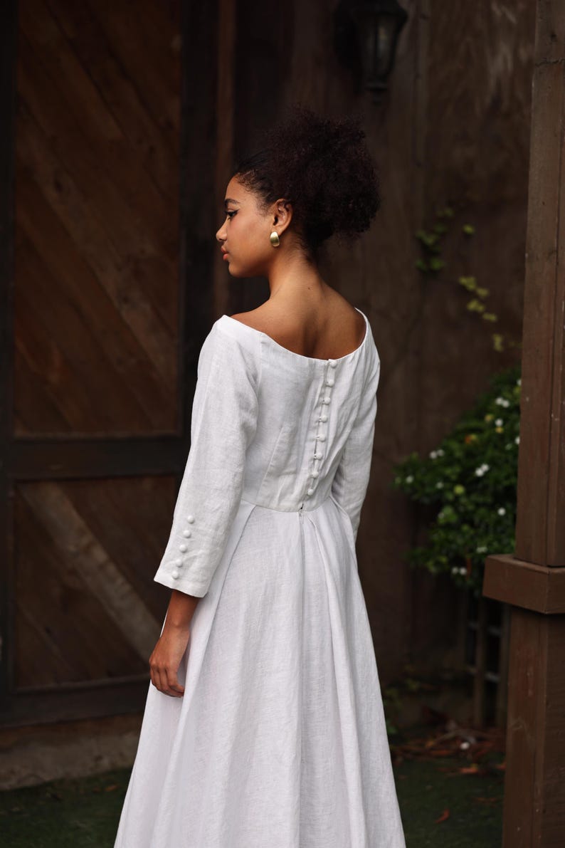 A close-up rear view of the model focusing on the upper back design. The image reveals a stunning row of meticulously handcrafted fabric-covered buttons lining the spine above an invisible zipper closure, adding a refined, classic bridal finish to the clean linen bodice.