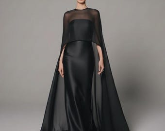 May include: A full-length image of a woman wearing a black evening gown with a sheer cape. The gown is floor-length with a fitted bodice and a flowing skirt. The cape drapes over the shoulders and arms, adding a dramatic flair to the formal attire.