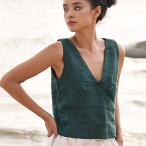 May include: A dark green sleeveless top with a deep V-neck. The top is made of a textured fabric and is paired with light-coloured trousers. The model is standing outdoors with the sea in the background.