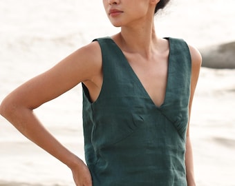 May include: A dark green sleeveless top with a deep V-neck. The top is made of a textured fabric and is paired with light-colored pants. The model is standing outdoors with the ocean in the background.