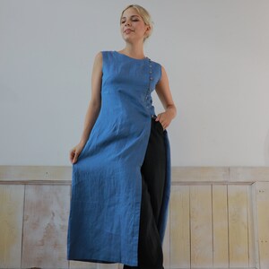 Full-length front view of the model standing poised in a studio setting, showcasing the long asymmetrical hemline and clean sleeveless design of the blue linen tunic garment.
