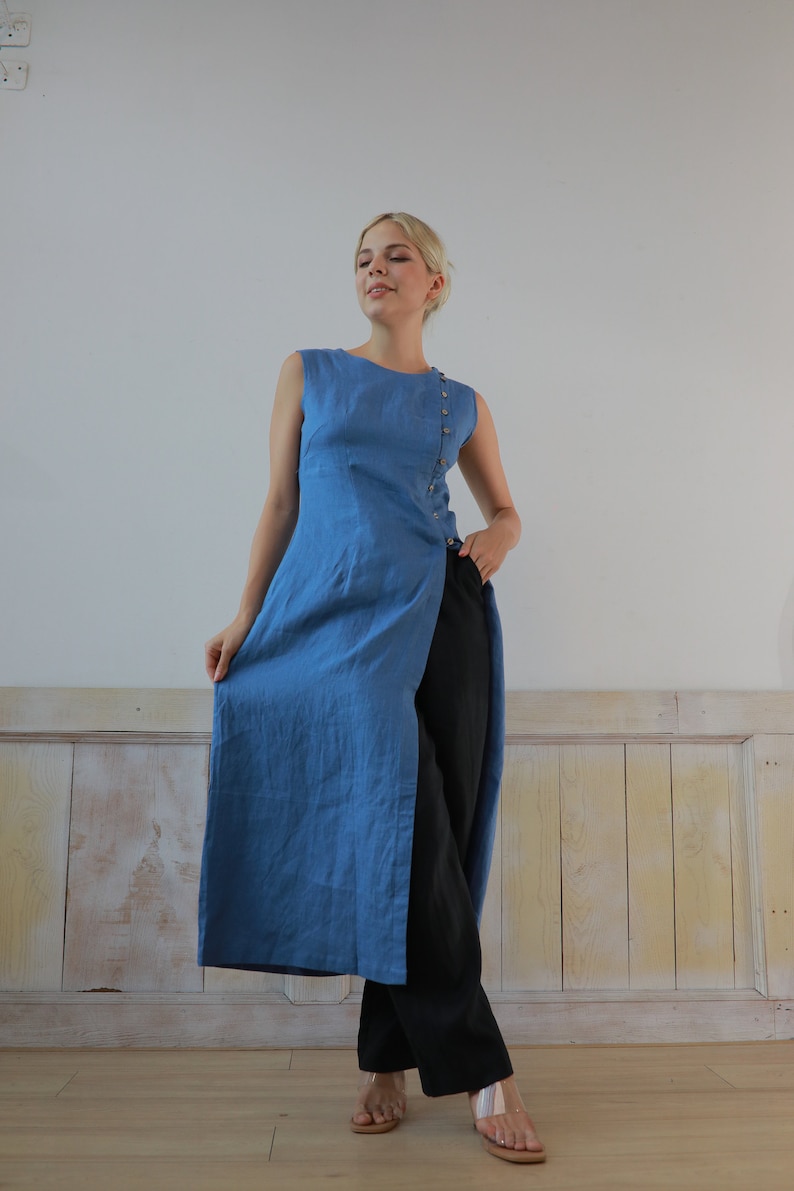 Full-length front view of the model standing poised in a studio setting, showcasing the long asymmetrical hemline and clean sleeveless design of the blue linen tunic garment.