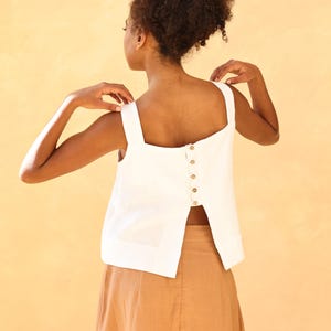 A upper-body rear profile of the model posing with her hands adjusting her wide shoulder straps against a warm beige background. This angle highlights the comfortable, relaxed fit and the functional timber-textured buttons along the back seam.
