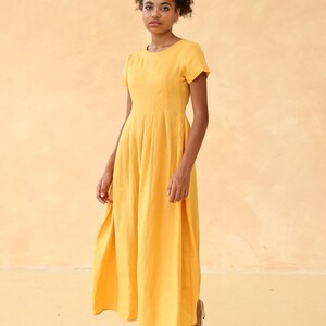 Full-length front view of a woman standing and looking slightly down, modeling a mustard yellow short-sleeved linen maxi dress, overlaid with editing interface icons.