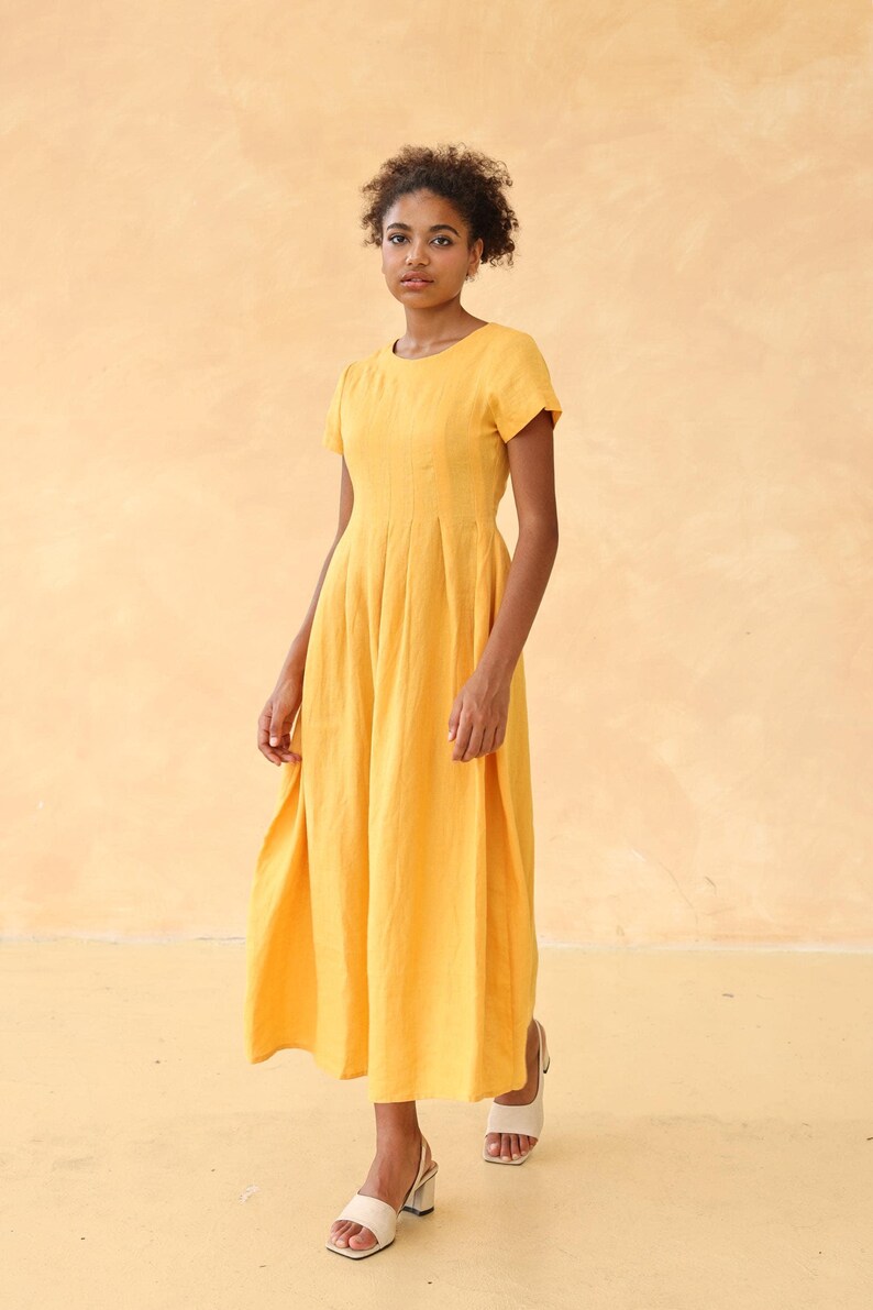 Full-length front view of a woman standing and looking slightly down, modeling a mustard yellow short-sleeved linen maxi dress, overlaid with editing interface icons.