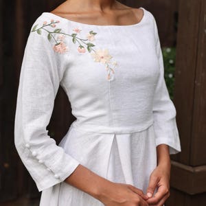 A close-up view of the dress's bodice on the model from a side-front angle. It highlights the delicate hand-embroidered floral branch cascading gently over the right shoulder and collarbone, the clean boat neckline, and the fine fabric-covered button details on the cuffs of the 3/4 sleeves.