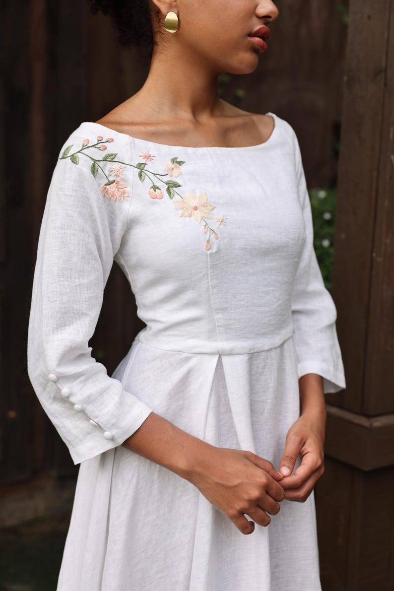 A close-up view of the dress's bodice on the model from a side-front angle. It highlights the delicate hand-embroidered floral branch cascading gently over the right shoulder and collarbone, the clean boat neckline, and the fine fabric-covered button details on the cuffs of the 3/4 sleeves.