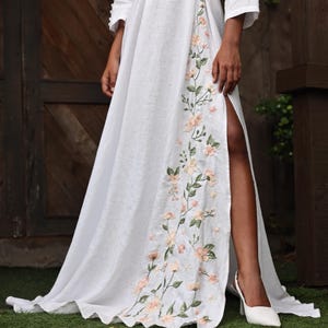 A close-up shot focusing on the lower half of the white linen dress, highlighting a high front slit that reveals the model's leg and white high heels. The edge of the slit and the flowing floor-length hem are adorned with intricate, hand-embroidered floral motifs in soft peach and green tones.
