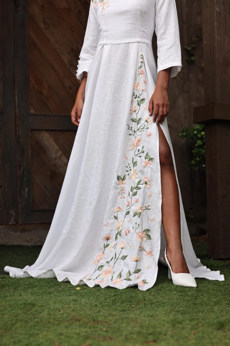 A close-up shot focusing on the lower half of the white linen dress, highlighting a high front slit that reveals the model's leg and white high heels. The edge of the slit and the flowing floor-length hem are adorned with intricate, hand-embroidered floral motifs in soft peach and green tones.
