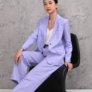 Full-length shot of a female model sitting on a black chair against a grey studio wall, wearing a two-piece lilac linen suit set featuring a single-breasted blazer and matching high-waisted wide-leg trousers.