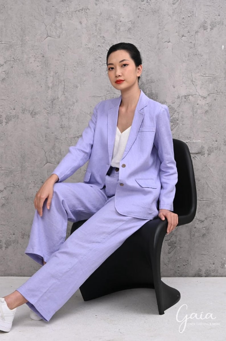 Full-length shot of a female model sitting on a black chair against a grey studio wall, wearing a two-piece lilac linen suit set featuring a single-breasted blazer and matching high-waisted wide-leg trousers.