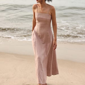 Full-length front view of a woman walking along the shoreline on a sandy beach, wearing a dusty pink linen maxi dress with vertical seam details.