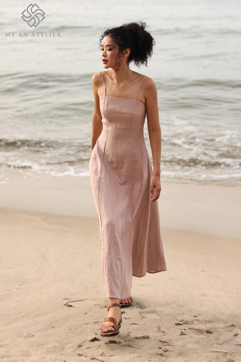 Full-length front view of a woman walking along the shoreline on a sandy beach, wearing a dusty pink linen maxi dress with vertical seam details.