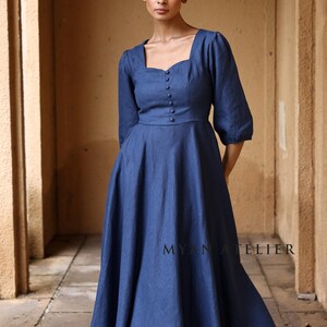Medium front view from the thighs up of a woman standing straight under a stone archway, modeling a classic indigo blue linen long dress with tailored panel seams and small fabric-covered buttons on the chest.