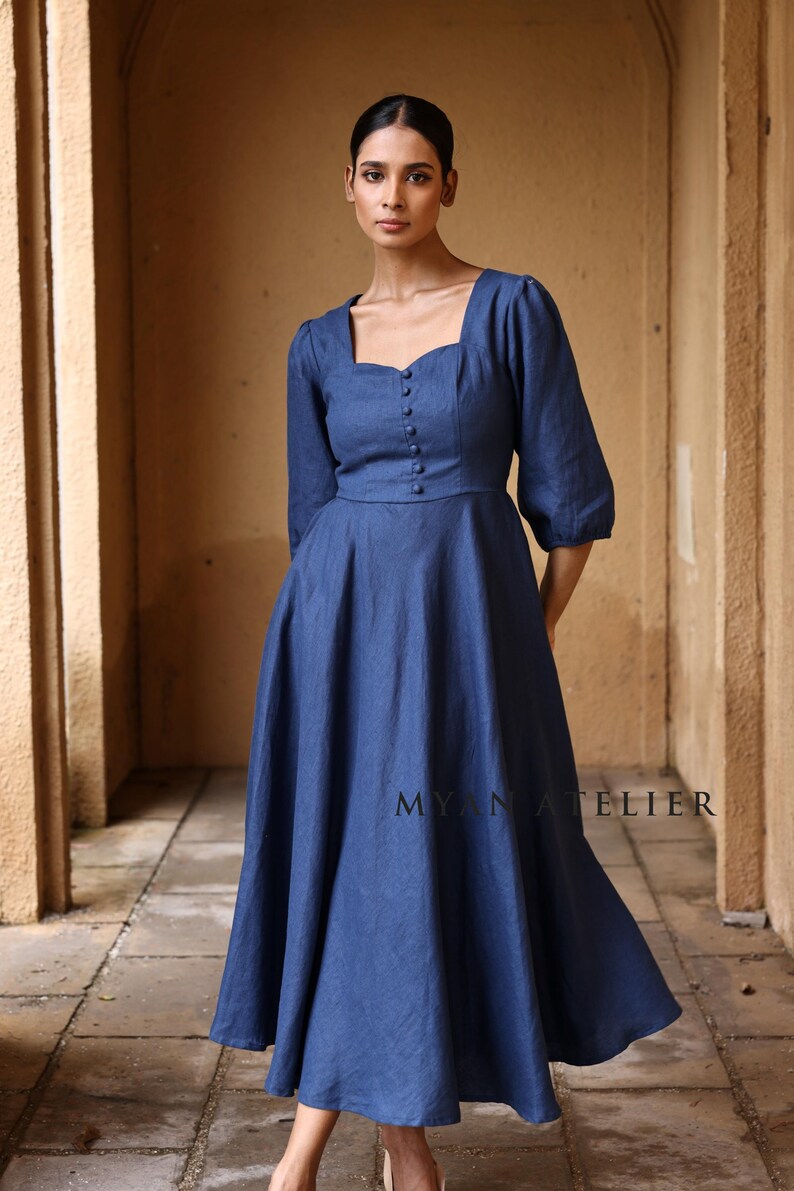 Medium front view from the thighs up of a woman standing straight under a stone archway, modeling a classic indigo blue linen long dress with tailored panel seams and small fabric-covered buttons on the chest.