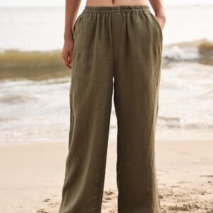 Full-length front view of the model standing with a relaxed posture on the sand, emphasizing the breezy, lightweight movement and casual summer holiday aesthetic of the olive green trousers.