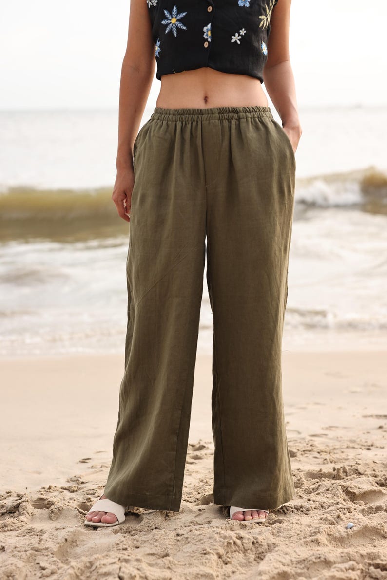 Full-length front view of the model standing with a relaxed posture on the sand, emphasizing the breezy, lightweight movement and casual summer holiday aesthetic of the olive green trousers.