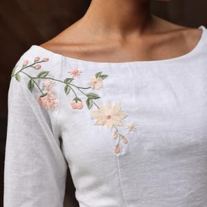 A tight close-up shot focusing on the wide boat neckline and chest area. This image perfectly highlights the harmony of the artisanal hand-embroidered floral bouquet on the right shoulder, emphasizing the high-quality craftsmanship and clean finishing of the neckline.