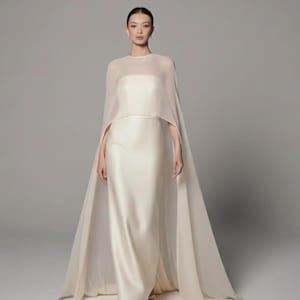 May include: A floor-length ivory satin gown with a sheer cape overlay. The cape drapes from the shoulders, creating a flowing silhouette. The dress has a simple, elegant design, suitable for formal events.