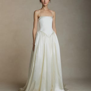 May include: A white bridal gown featuring a square neckline and delicate spaghetti straps. The fitted bodice flows into a full, A-line skirt. The dress is crafted from a smooth, satin-like material, creating an elegant look.