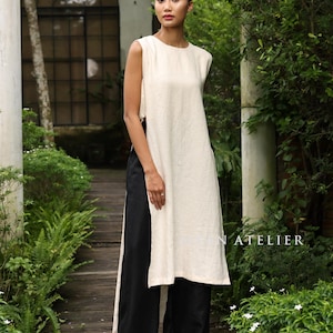 May include: A cream sleeveless tunic and black wide-leg trousers. The tunic has a high neckline and a side slit. The trousers are floor-length. The background is a garden with greenery and a wooden walkway. The text "N ATELIER" is visible.