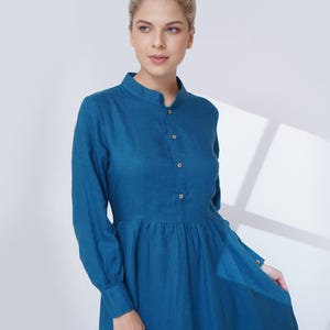 May include: A teal dress with a mandarin collar and long sleeves. The dress has a button-down front and a gathered waist, creating a flowing shape. The fabric looks like linen or a similar lightweight material, suitable for warmer weather.