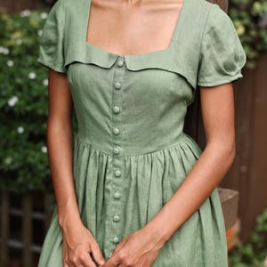 May include: A sage green dress with a square neckline and short, puffed sleeves. The dress features a button-down front and a fitted bodice that flows into a pleated skirt. The fabric appears to be linen or a similar lightweight material, suitable for warm weather.