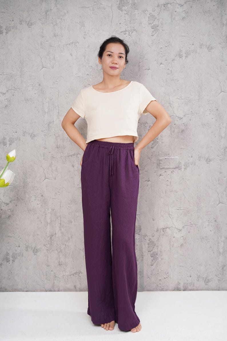 Full-length front shot of a model standing against a grey studio wall, wearing loose high-waisted magenta purple linen wide-leg trousers with an elasticated waistband, paired with a short-sleeve off-white top.