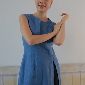 A close-up front view from the waist up of the model with her hands raised near her chest, highlighting the round neckline and the delicate side button detailing along the bodice of the tunic.