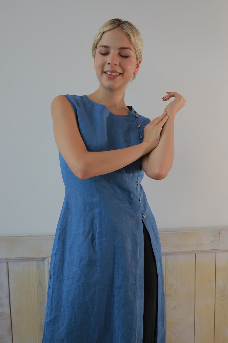 A close-up front view from the waist up of the model with her hands raised near her chest, highlighting the round neckline and the delicate side button detailing along the bodice of the tunic.