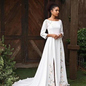 A full-length shot of a model standing outdoors against a dark wooden structure, wearing the white linen A-line wedding dress. The photo showcases its elegant boat neckline, structured 3/4 sleeves, a fitted waist with soft pleats, and a full-volume skirt featuring a high front slit and a subtle train pooling on the grass.