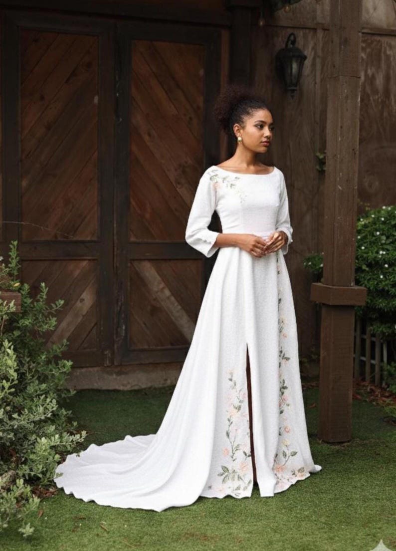 A full-length shot of a model standing outdoors against a dark wooden structure, wearing the white linen A-line wedding dress. The photo showcases its elegant boat neckline, structured 3/4 sleeves, a fitted waist with soft pleats, and a full-volume skirt featuring a high front slit and a subtle train pooling on the grass.