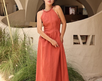 May include: A rust-colored halter-neck maxi dress with a braided neckline. The dress has a fitted bodice and a flowing skirt. The model is wearing white sandals. The dress is from MYAN ATELIER.
