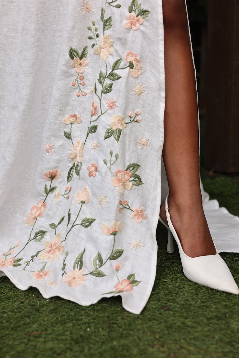A detailed close-up shot focusing on the exquisite hand-embroidery along the dress's front slit. It showcases the delicate needlework stitches and rich texture of the embroidery floss forming a climbing vine of peach-colored blossoms and green leaves on the softened white linen.