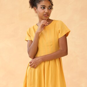 Medium front shot from the waist up of a woman with a thoughtful expression, wearing a mustard yellow linen dress with short cap sleeves against a peach background.