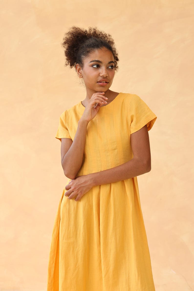 Medium front shot from the waist up of a woman with a thoughtful expression, wearing a mustard yellow linen dress with short cap sleeves against a peach background.
