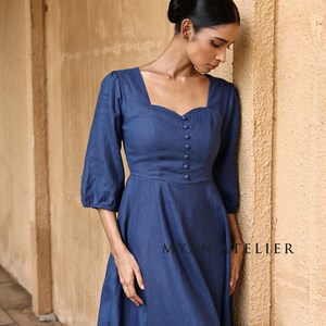 Medium front three-quarter view of a woman leaning thoughtfully against a wall, focusing on the structured bodice panels and retro cottagecore charm of a slate blue three-quarter sleeved linen maxi dress.