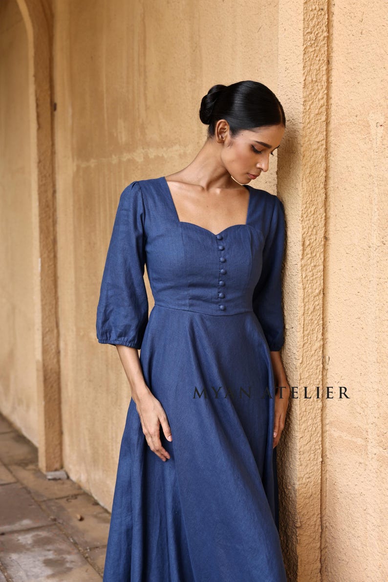 Medium front three-quarter view of a woman leaning thoughtfully against a wall, focusing on the structured bodice panels and retro cottagecore charm of a slate blue three-quarter sleeved linen maxi dress.