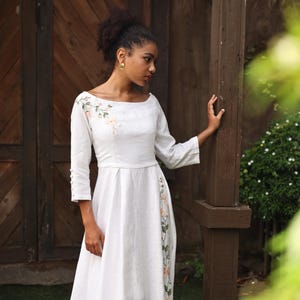 A full-length shot of the model posing with one hand resting on a wooden pillar. The angle highlights the regular fit and elegant profile of the white embroidered linen gown, showing the balance between the simple sleeveless-looking front silhouette and the detailed side slit.