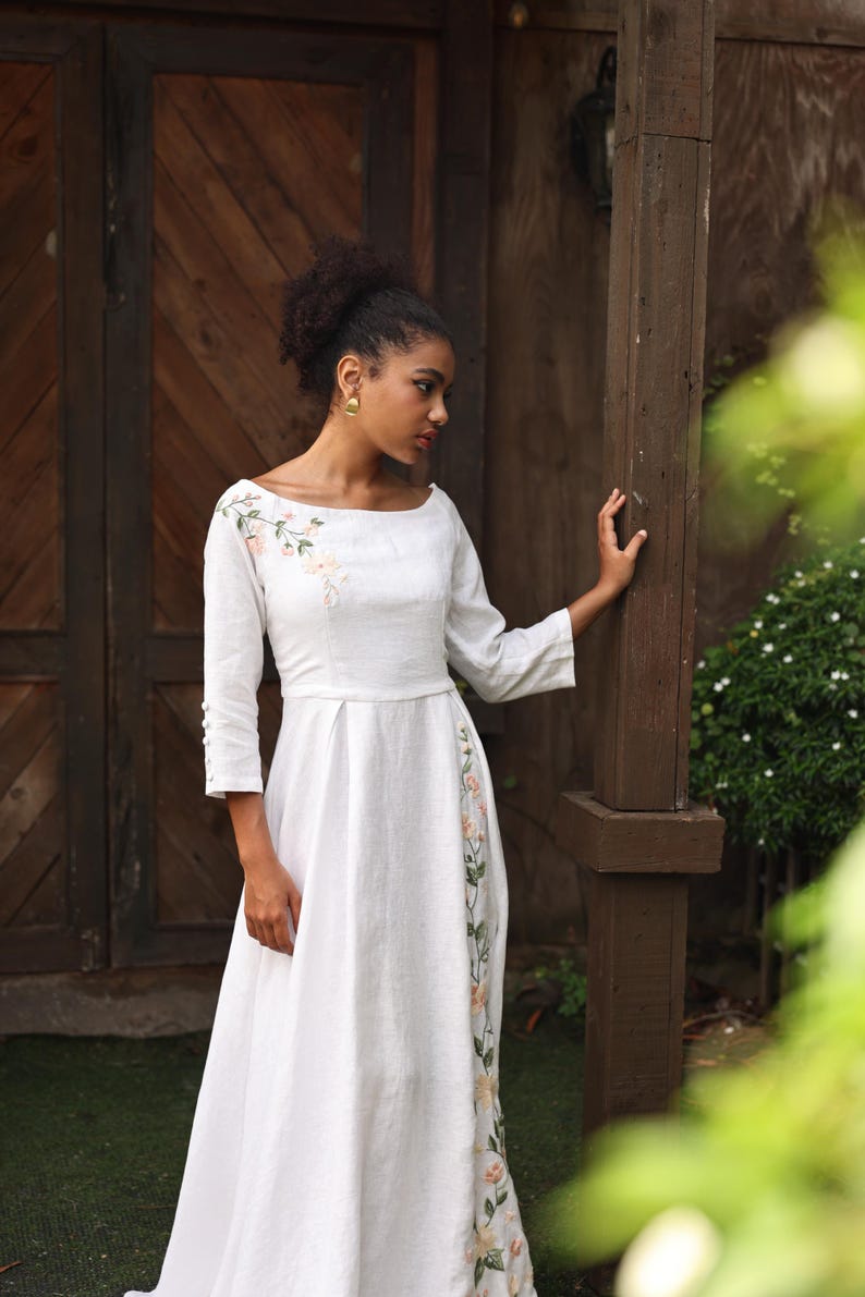 A full-length shot of the model posing with one hand resting on a wooden pillar. The angle highlights the regular fit and elegant profile of the white embroidered linen gown, showing the balance between the simple sleeveless-looking front silhouette and the detailed side slit.