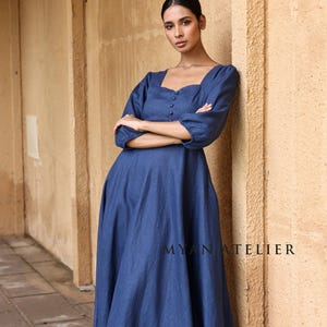 Full-length front shot of a woman leaning with crossed arms against a textured pillar, showcasing the relaxed empire waistline and fluid drape of a slate blue linen maxi dress.