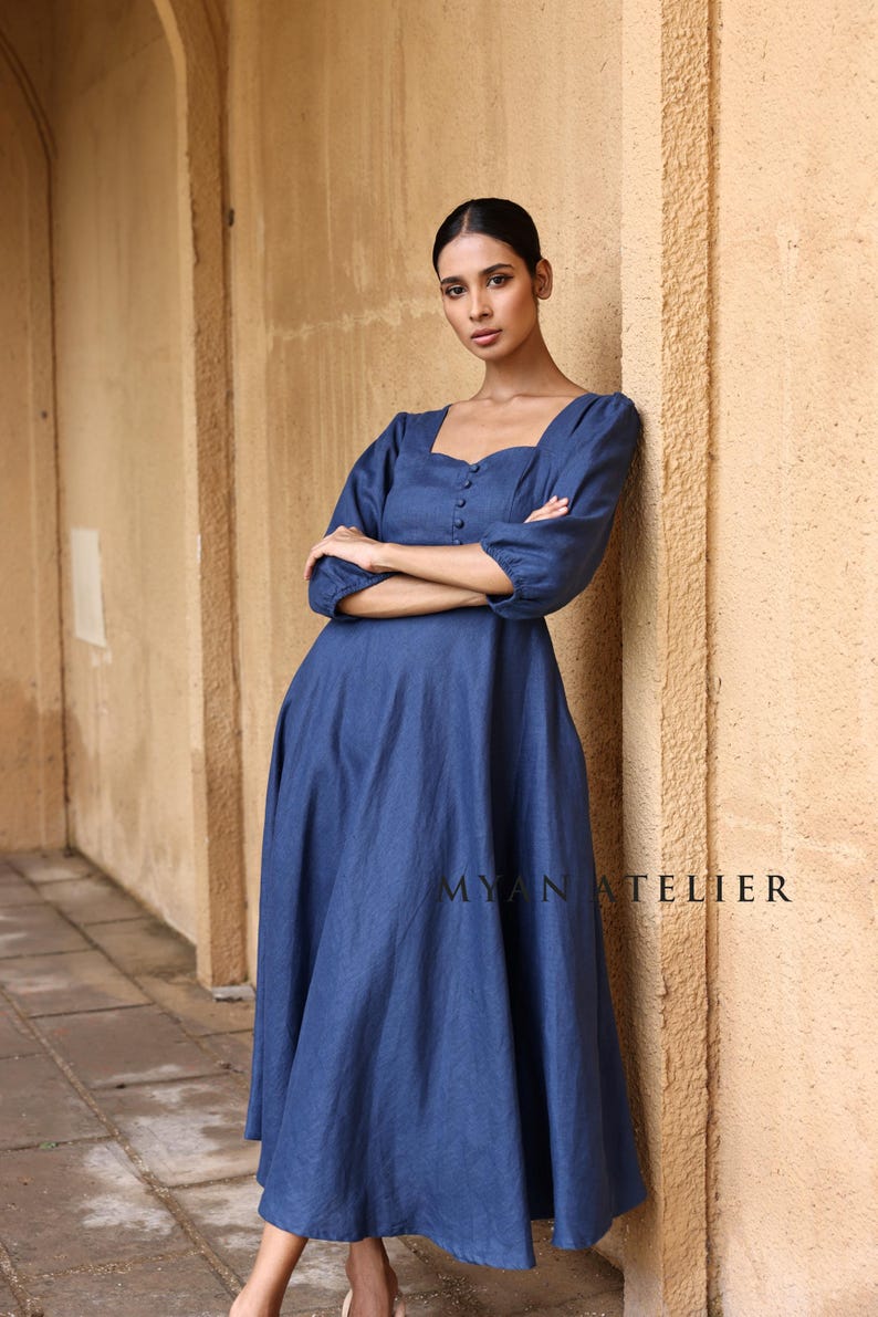 Full-length front shot of a woman leaning with crossed arms against a textured pillar, showcasing the relaxed empire waistline and fluid drape of a slate blue linen maxi dress.