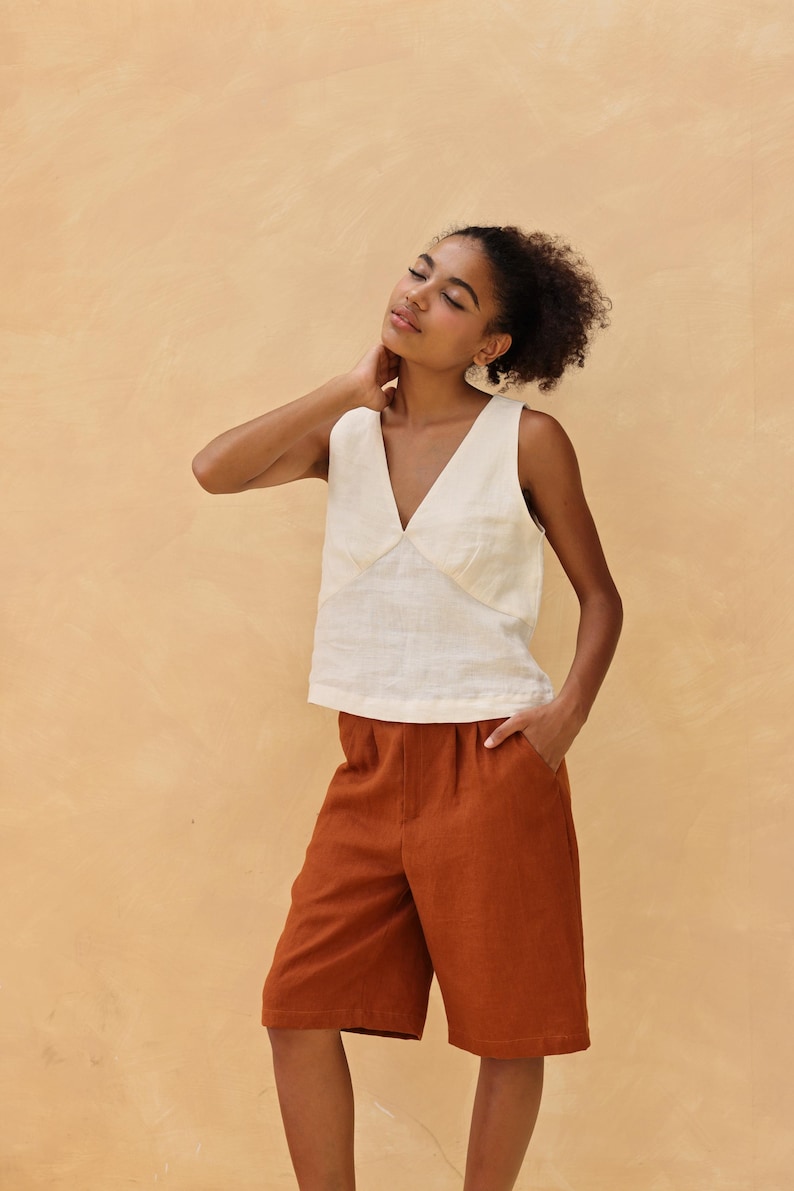 Medium front shot of the model leaning her head back with a relaxed expression against a warm beige wall, showcasing the casual cropped fit and lightweight texture of the cream linen vest top.