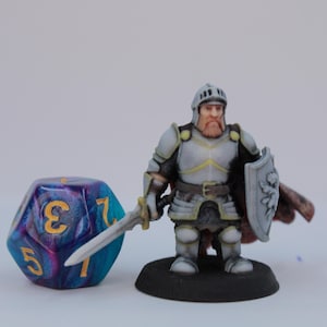 Könnte beinhalten: Eine Miniatur-Ritterfigur mit Schwert und Schild steht neben einem großen, mehrfarbigen, zwanzigseitigen Würfel. Der Ritter ist in silberner Rüstung und einem Helm gekleidet, mit einem roten Umhang. Der Würfel ist blau, lila und gold.